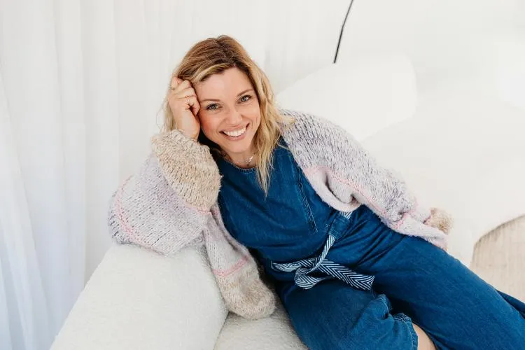 laura fox chronic illness mums business coach on sofa looking at camera in blue denim jumpsuit with pastel pink and purple cardigan