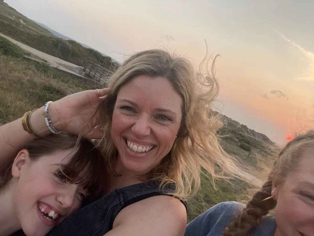 i am Laura business coach for ambitious women with health conditions. Laura and her 2 children, laughing at the camera s the wind blow their hair with the backdrop of a sunset at the beach.