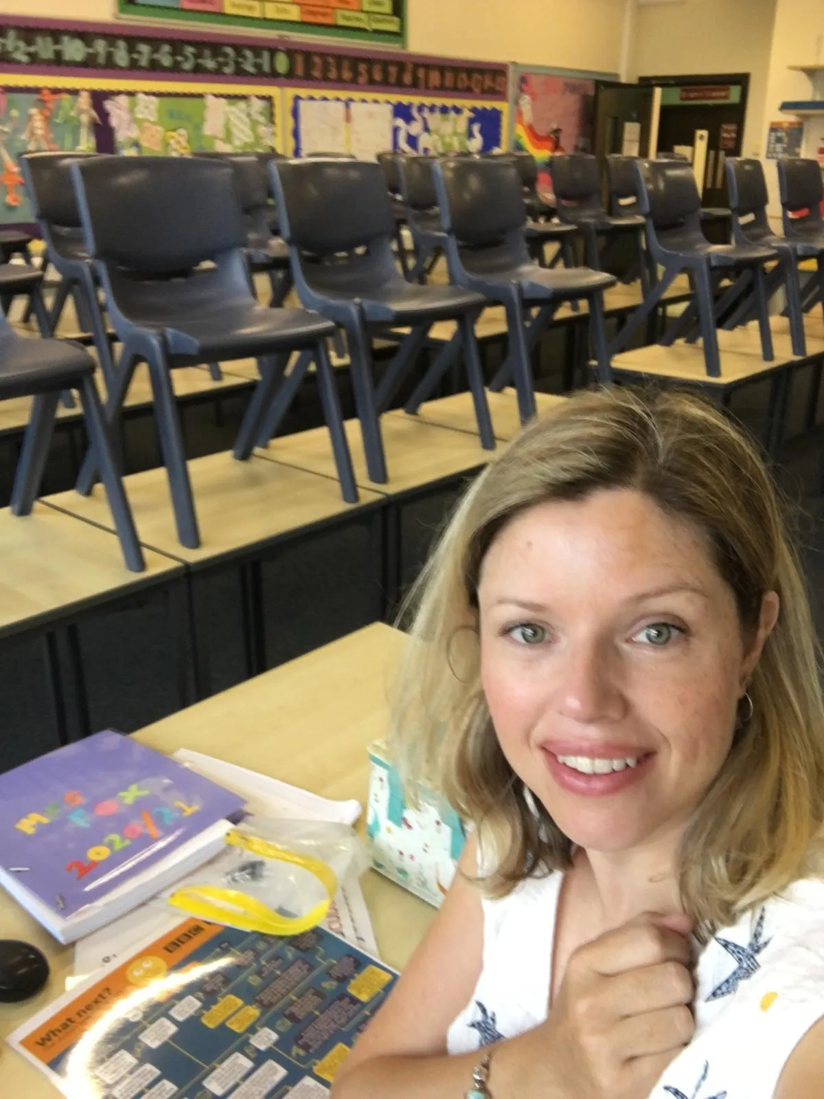 i am Laura business coach for ambitious women with health conditions. Laura sat in her old classroom, smiling at the camera, chairs on the desks behind her.