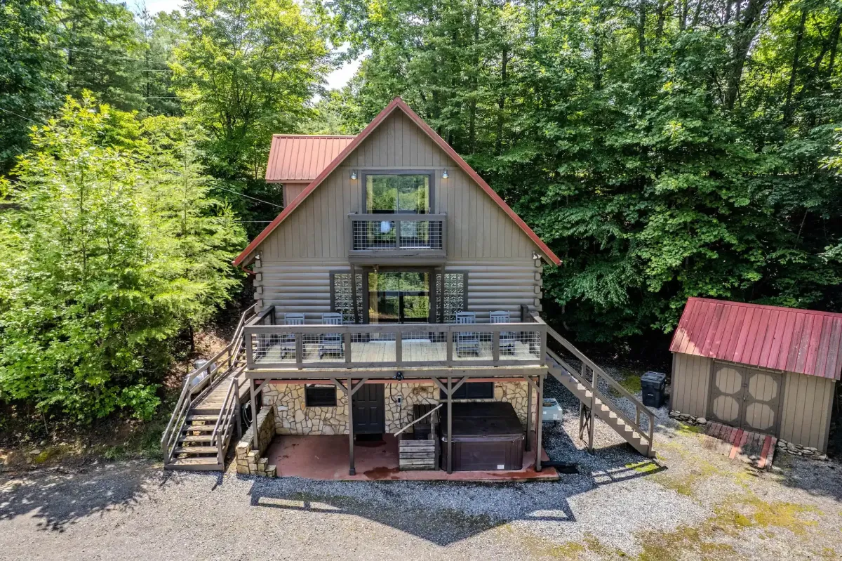 Rustic two-story log cabin with red metal roof and wraparound porch, nestled in a lush forest setting. The cabin features a spacious upper deck with modern railings and outdoor seating, a lower-level patio with a hot tub, and a small adjacent shed with matching red roof. Surrounded by dense green trees, the property sits on a gravel clearing ideal for mountain retreats.