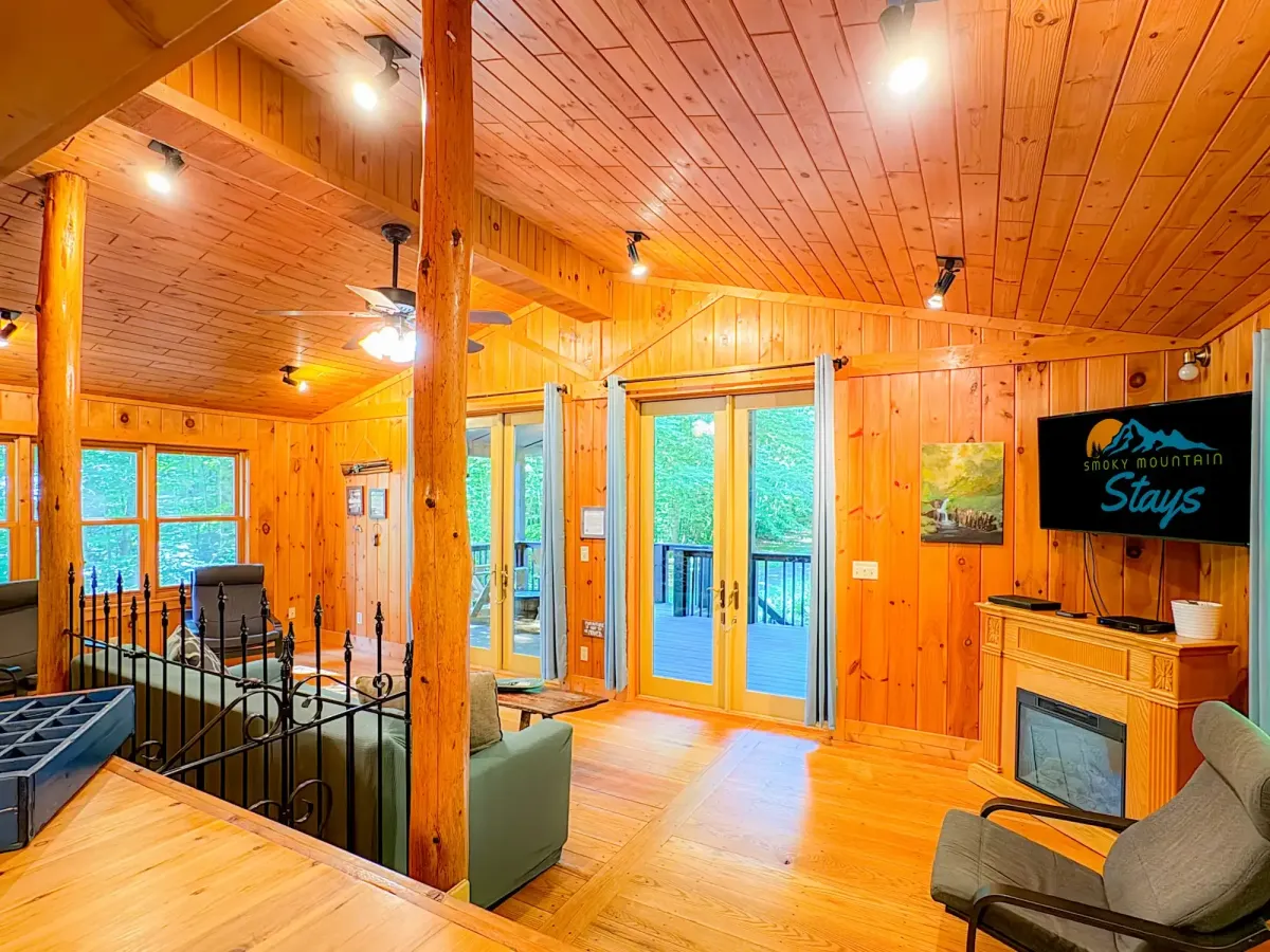 Cozy cabin interior with polished pine walls, ceilings, and floors, showcasing a warm, rustic ambiance. The open living area includes modern lounge chairs, a faux fireplace with a mounted flat-screen TV displaying "Smoky Mountain Stays," and large French doors leading to a forest-view deck. Exposed log beams and track lighting enhance the natural wood aesthetic, while large windows bring in abundant daylight and scenic greenery.