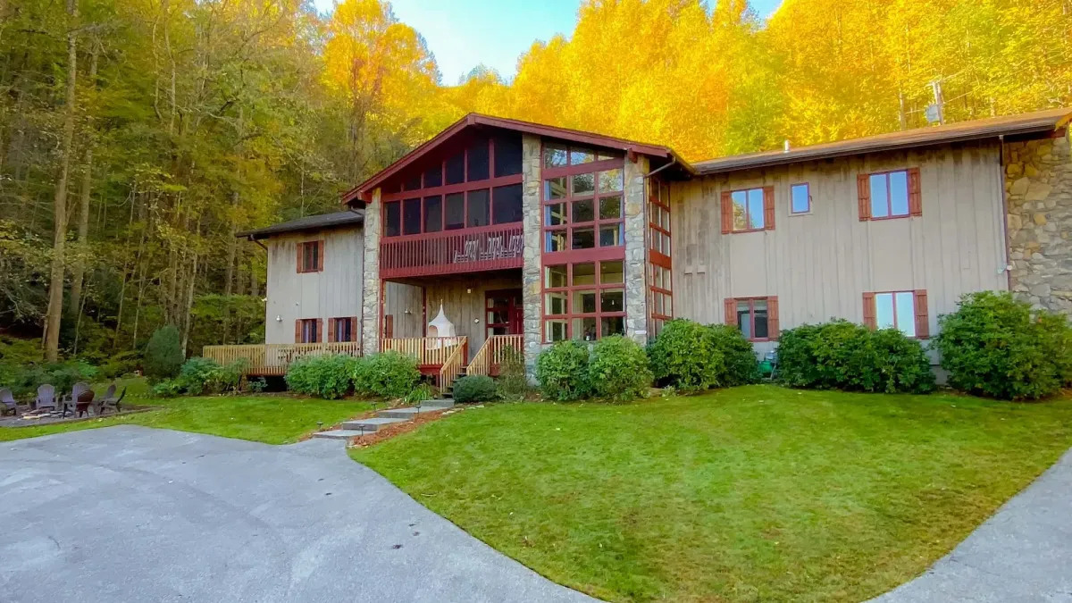 Expansive mountain lodge-style home with rustic wood siding, stone accents, and large floor-to-ceiling windows centered in a dramatic A-frame entrance. The structure features a second-story screened-in balcony, multiple gables, and charming red trim. Surrounded by manicured shrubs and lawn, the home is nestled at the edge of a vibrant autumn forest, with golden foliage glowing behind. A curved driveway and outdoor fire pit area complete the inviting mountain retreat setting.