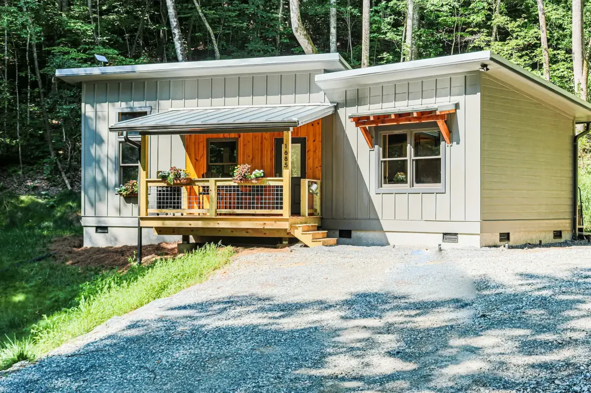 Modern cabin with a sleek, minimalist design featuring vertical board-and-batten siding in a soft gray tone, accented by warm natural wood trim. A small covered front porch with wire railing, wooden posts, and colorful flower boxes adds charm to the entrance. Set against a forested backdrop, the cabin sits on a gravel driveway and offers a peaceful, woodland retreat vibe with contemporary rustic appeal.