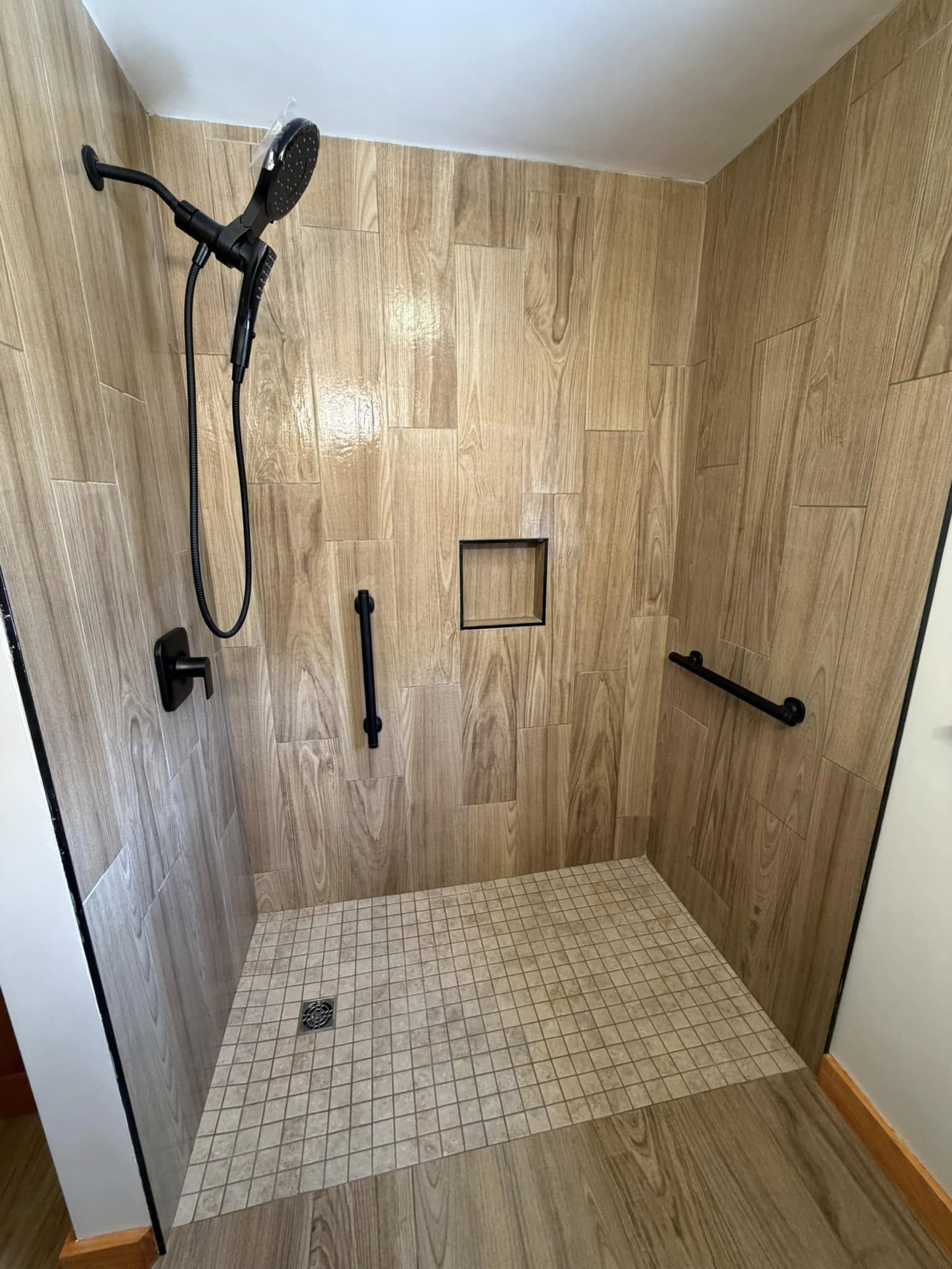 Custom tile shower installation by Grams Tile — large format wood-look porcelain wall tile, matte black fixtures, grab bar, recessed niche, and small format mosaic floor