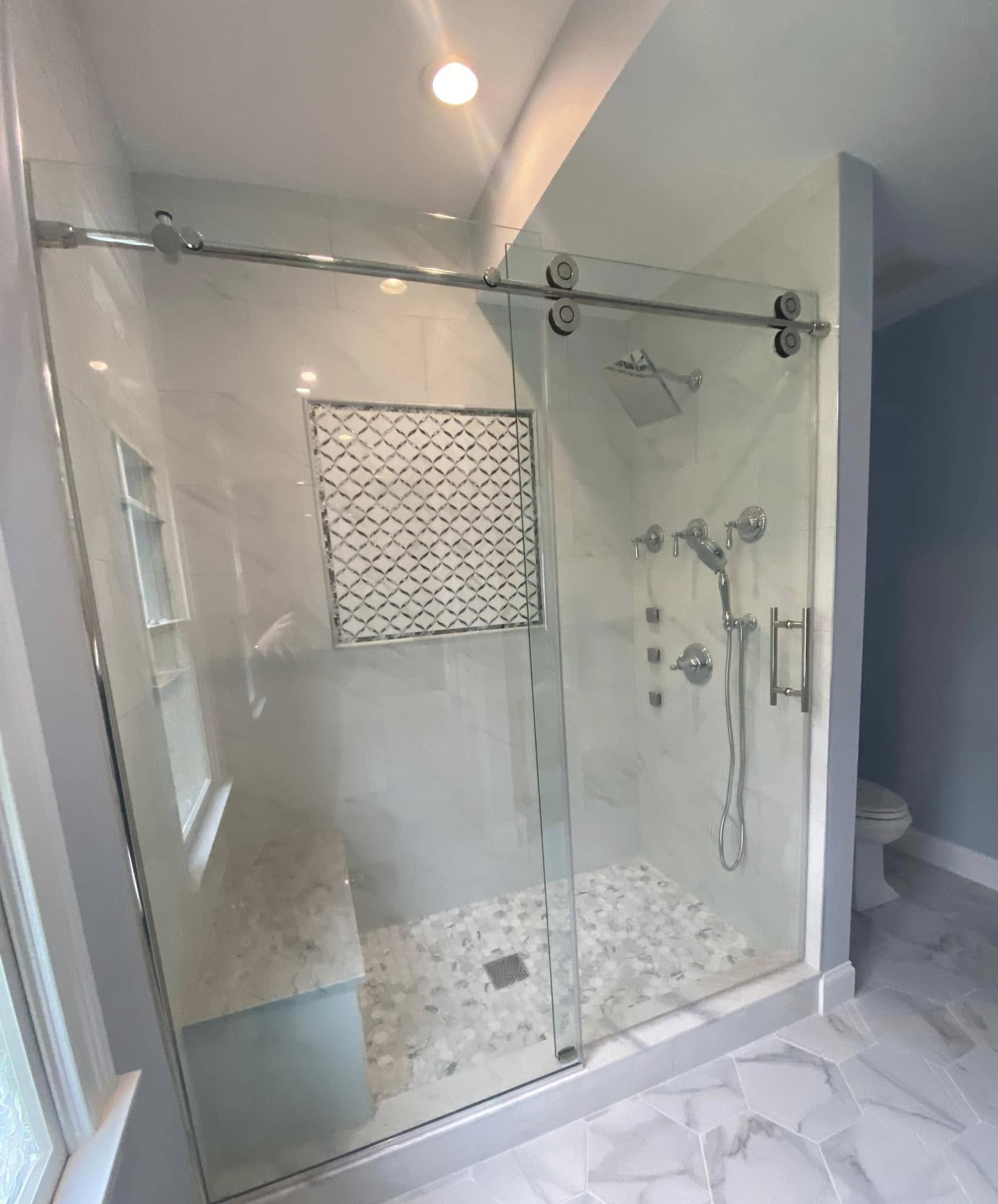 Walk-in shower remodel by Grams Tile in Lunenburg MA — large format light gray porcelain tile, sliding frameless glass barn door, decorative Moroccan tile niche, and pebble mosaic floor