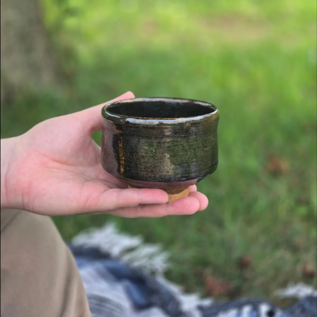 A handmade black tea bowl resting in an open hand against a softly blurred green garden.