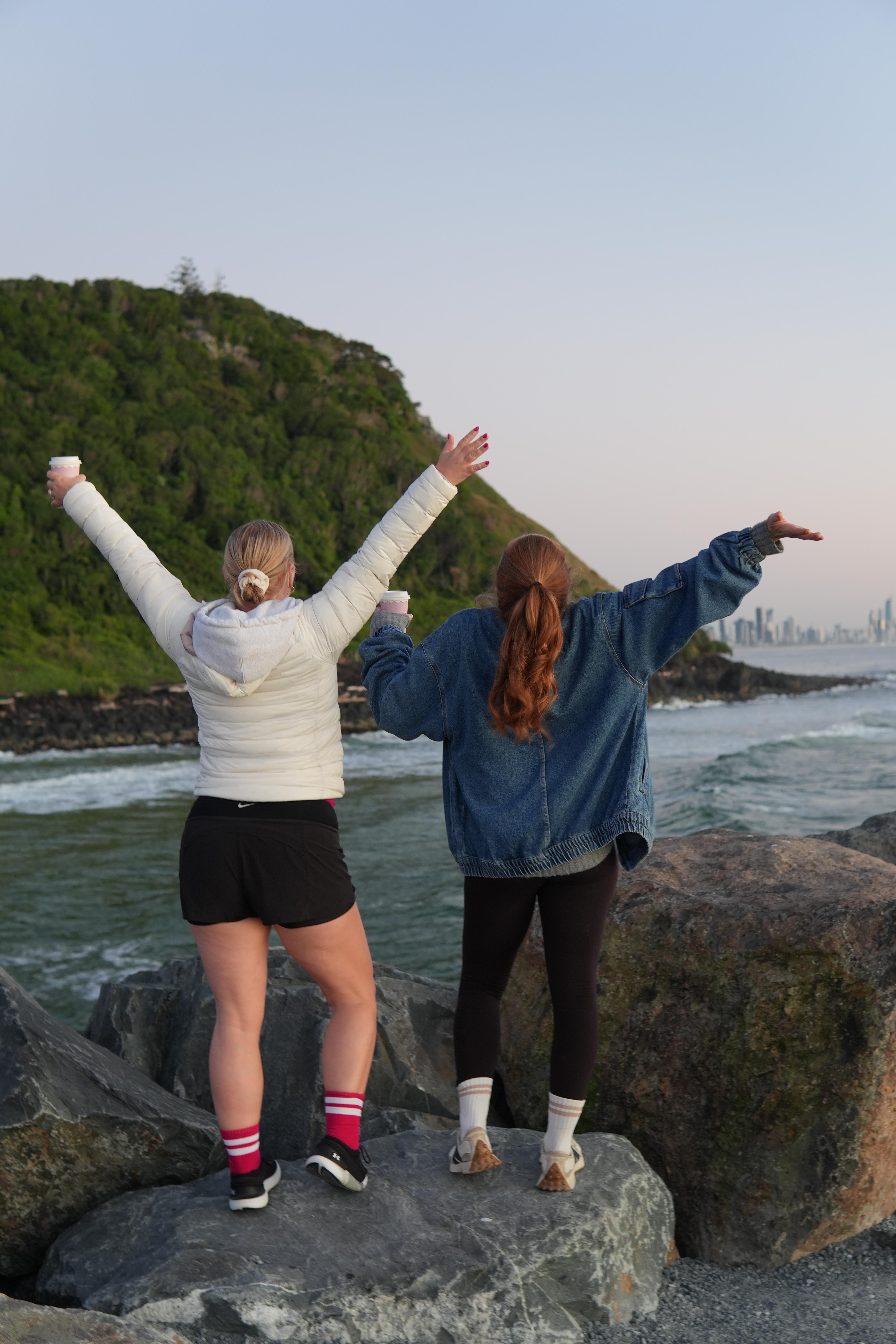 Photo of two people standing on rocks at the beach with their hands raised looking out to the water