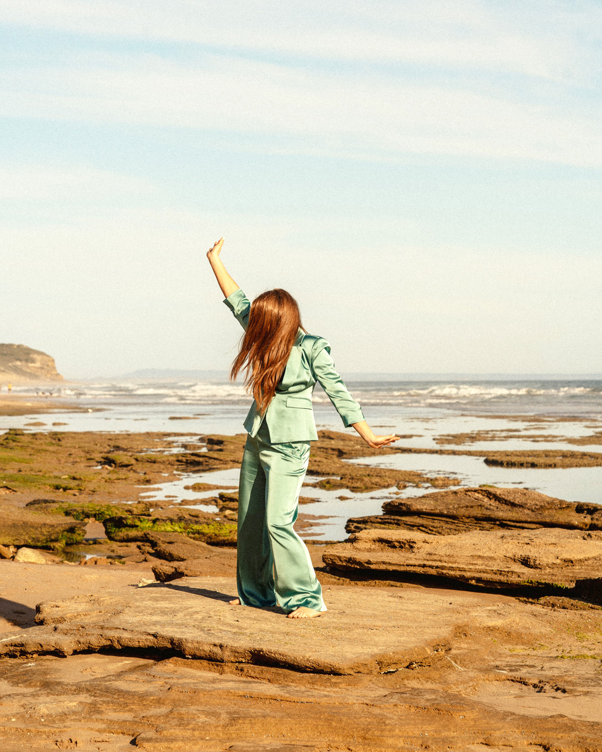 Marejka Knigge - Lady standing on rock at the beach, with ocean view in the background,  wearing aqua green suit and red flowing hair. Manifestation Coach, Life Coach.
