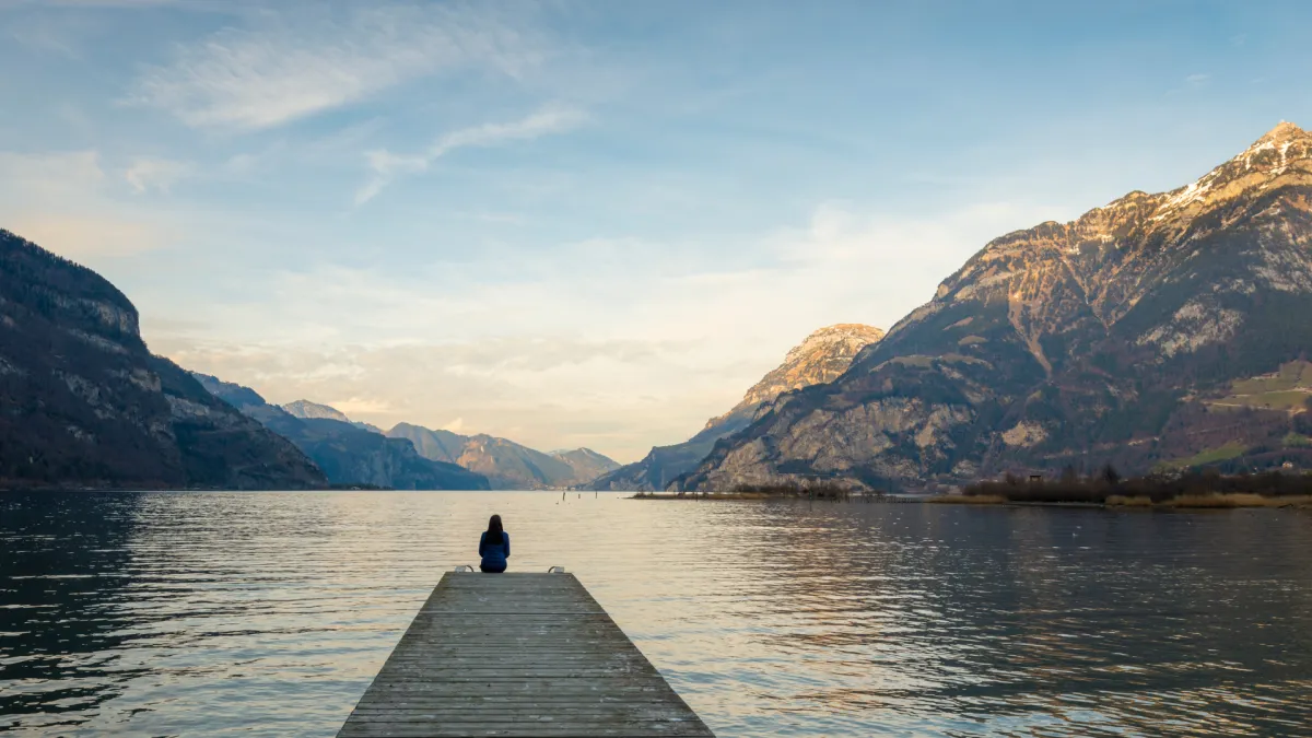 Person sitting on a wooden dock overlooking a peaceful mountain lake at sunset.