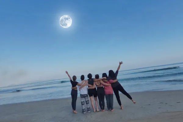 Six women embracing on a peaceful beach at sunset, looking toward the ocean under a full moon, representing community, transformation, and healing.