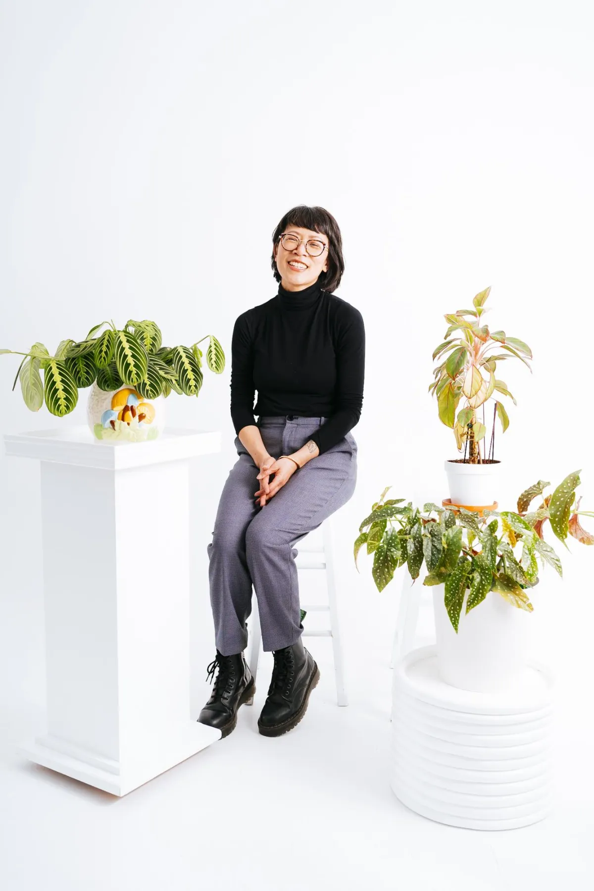 Photo of Xuan-Yen Cao in a black turtleneck, gray pants and black shoes, sitting on a white stool in a white room, surrounded by 3 potted plants