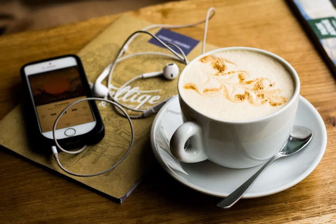 Latte in a cup and saucer, next to a mobile phone with wired earplugs, on a wooden table