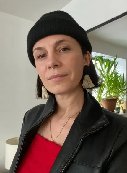 Photo of Aga Forfa wearing a red top, black jacket, black hat, gold necklace and tassel earrings, in a white room with plants in the background