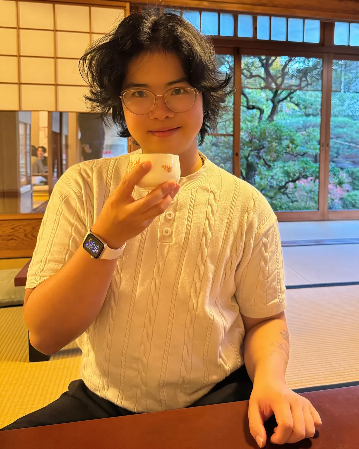 Photo of Yijia Shao wearing a white knit sweater, smartwatch and glasses, holding a white cup of tea, in a Japanese-style room with a lush garden in the background