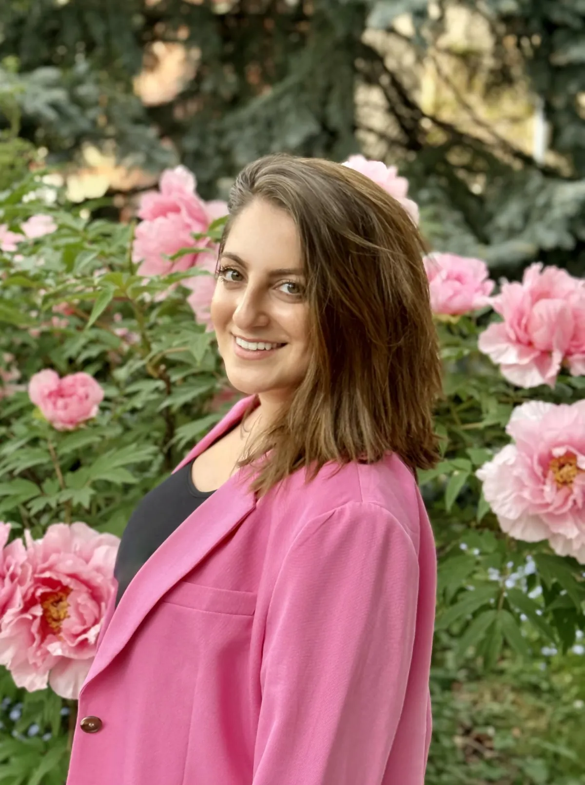Photo of Nikoo Sedaghat in black shirt and pink blazer, standing in front of a bush of pink flowers