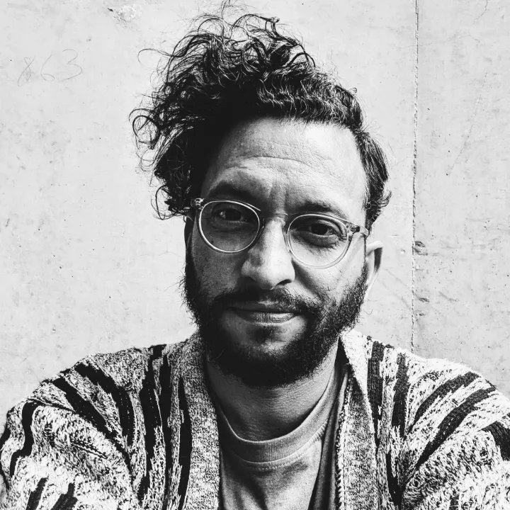 Black-and-white photo of Christopher McKinnon wearing a t-shirt and cardigan with glasses, on a blank grey background