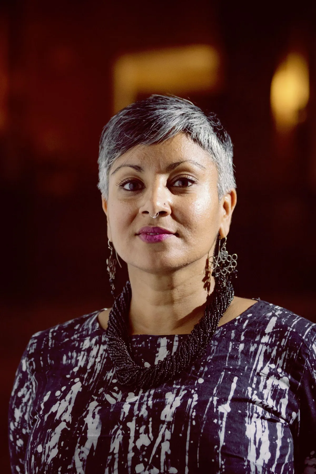 Photo of Tracey Jastinder Mann in a black and white top wearing a dark chunky necklace and drop-down earrings, in front of a blurry copper brown background