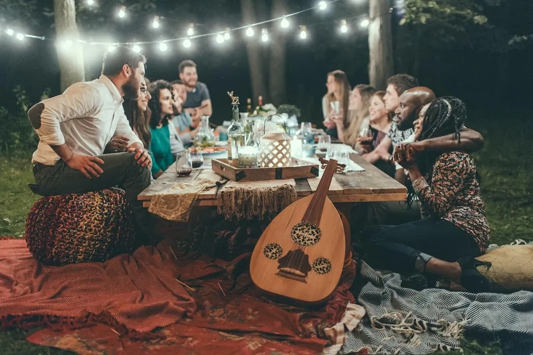 Diverse group of people from different cultural and racial backgrounds and of different genders sitting around a picnic table outdoors at night, enjoying a meal, beverages and conversation together, with a string of lights overhead, picnic blankets on the ground around them, and a guitar featured in the front-centre of the photo.
