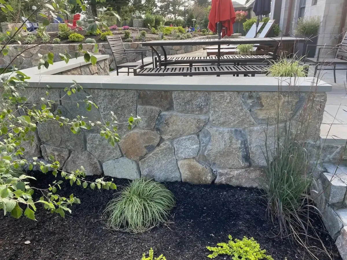 Natural stone masonry retaining wall with granite capstone and landscaped garden bed in Marblehead MA outdoor living space.