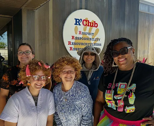 Five smiling R'Club staff members in playful costumes pose in front of the R'Club sign.
