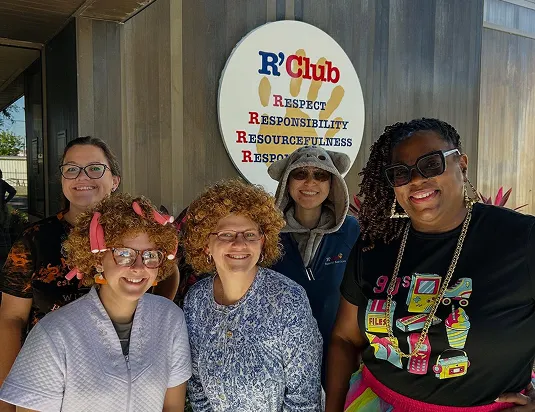 Five smiling R'Club staff members in playful costumes pose in front of the R'Club sign.