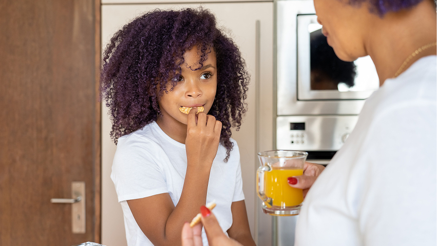 Child enjoying a healthy snack while an adult prepares food in the kitchen.