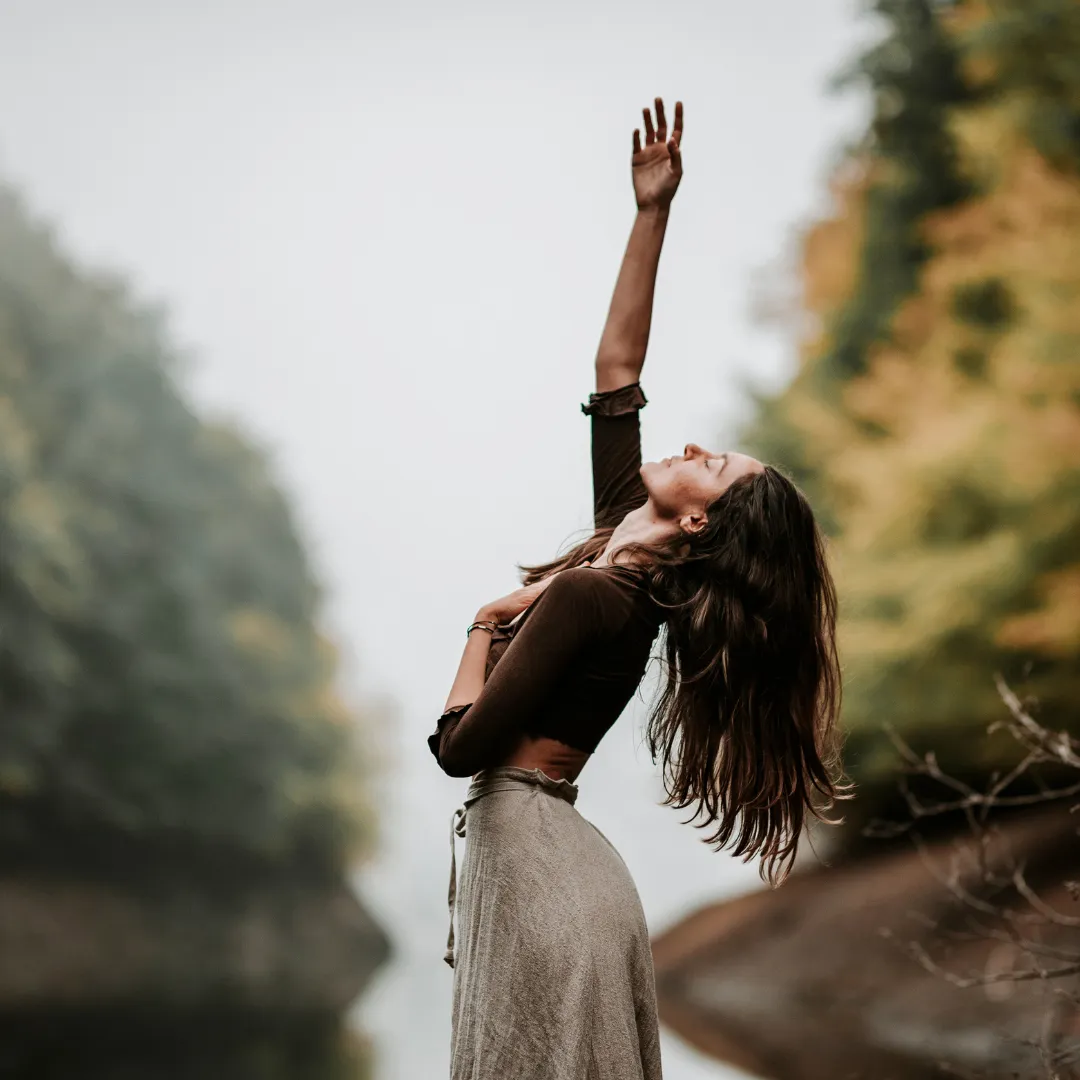 Woman in nature with one arm up and the other on her chest