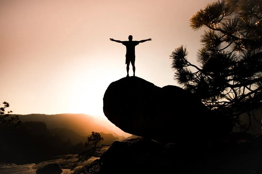 man standing on a bouder, arms out, sunset