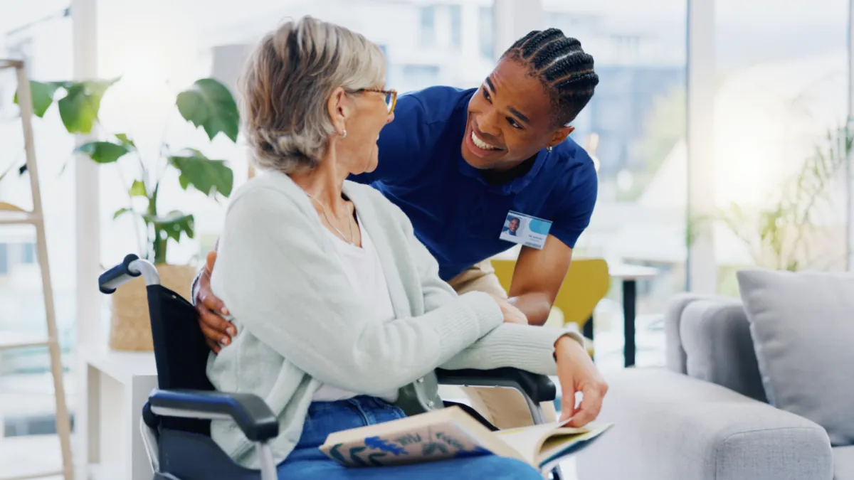 A cheerful male caregiver sits beside an elderly woman in a wheelchair, sharing a warm moment in a sunlit living room—representing NuLook Home Care’s commitment to dignity, connection, and supportive in-home services.