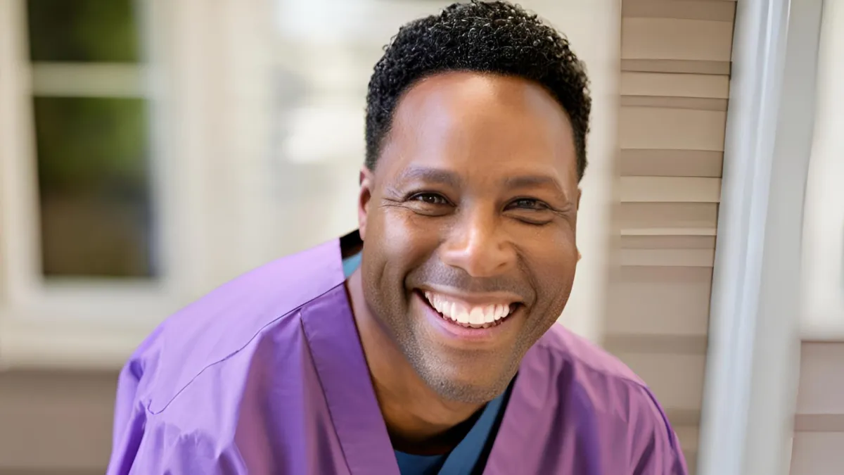 African American man smiling, with purple scrubs that have NuLook Home Care Agency written on the shirt