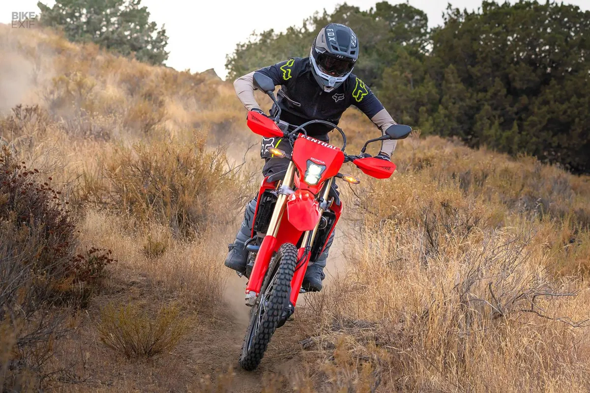 A motorcyclist in black gear riding a red off-road dirt bike uphill on a dry, grassy trail.