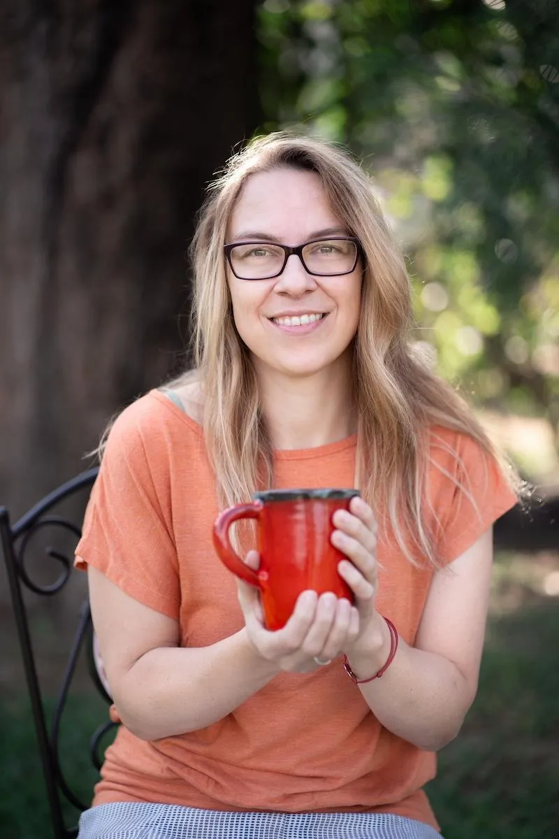 Eileen Ranscht smiling warmly, holding a red mug with both hands, seated outdoors — inviting calm and connection