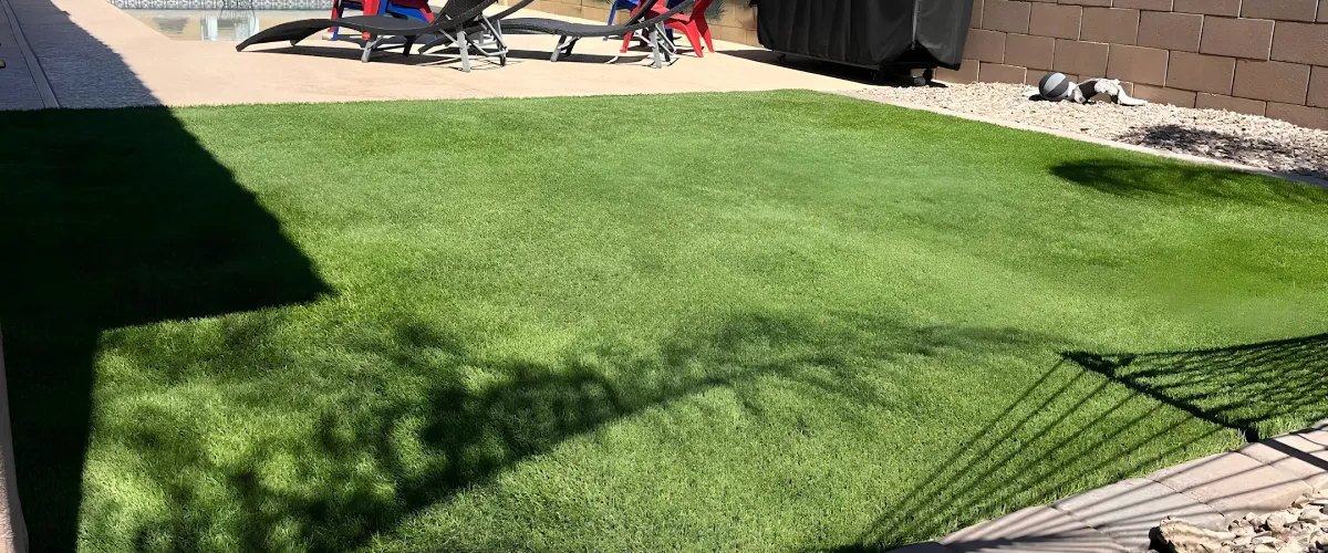 A backyard featuring green artificial turf with concrete paving, lounge chairs, and a pool area in the background.