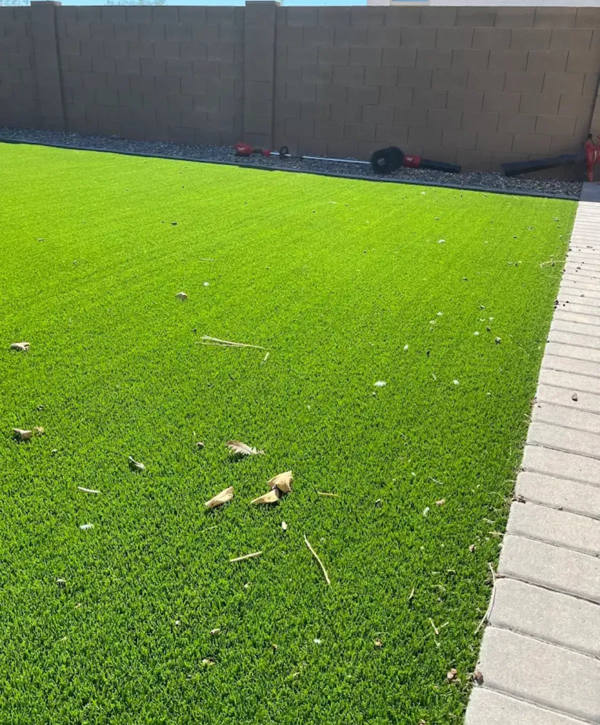 A bright green artificial turf backyard with a brown block wall and landscaping tools in the background.