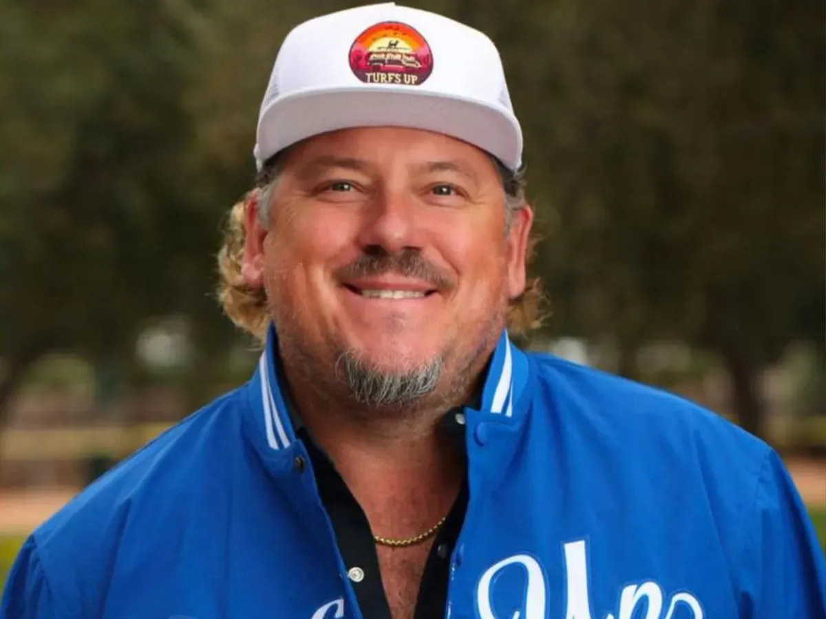 A smiling man with a beard and mustache wearing a white hat that says 'TURFS UP' and a blue jacket.