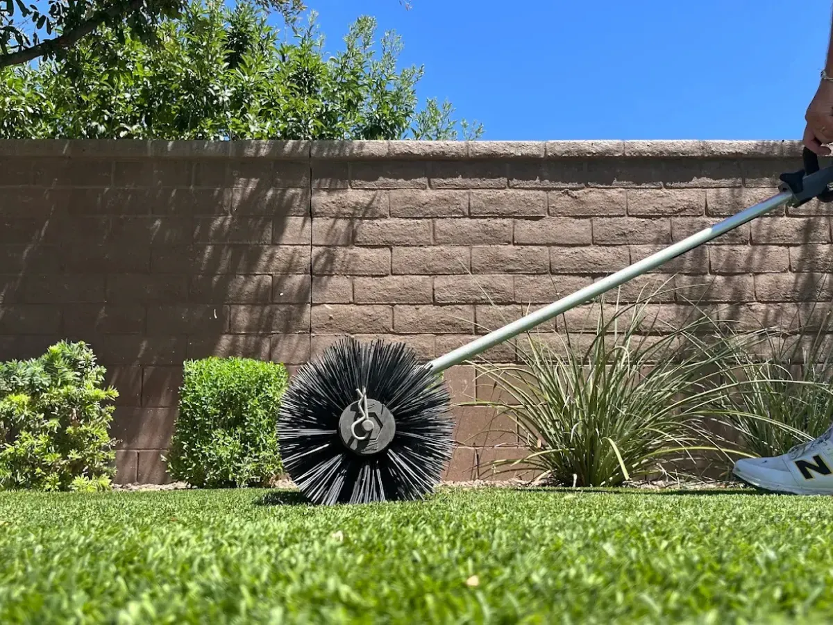 A large, black circular brush on a long handle grooming green artificial turf in a sunny backyard.