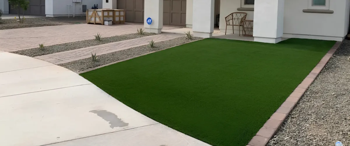 A suburban front yard featuring a bright green artificial turf lawn, a light-colored concrete pathway, and a paver driveway.