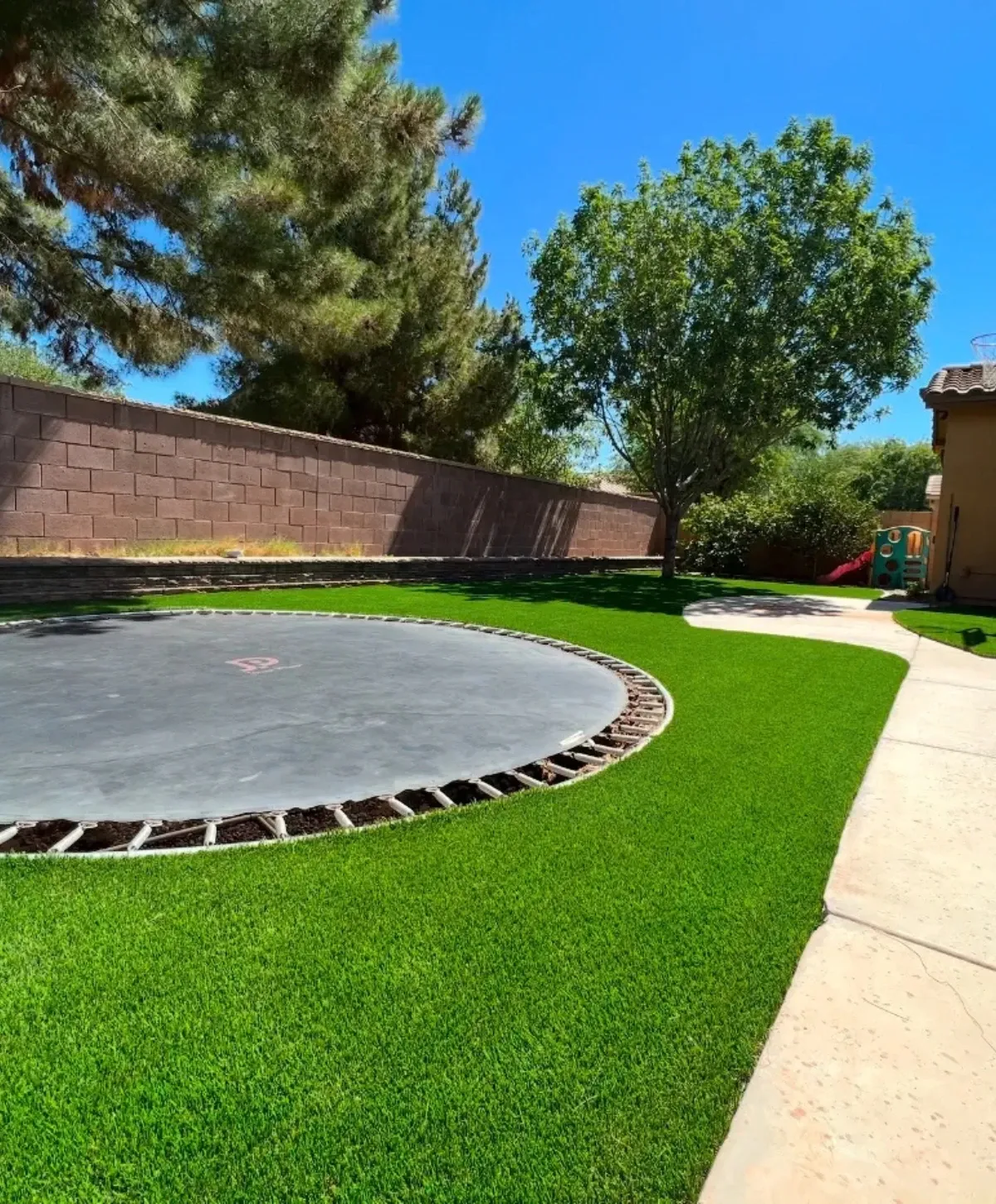 A backyard with an in-ground trampoline, vibrant artificial turf, a concrete path, and a brick wall under a sunny blue sky.