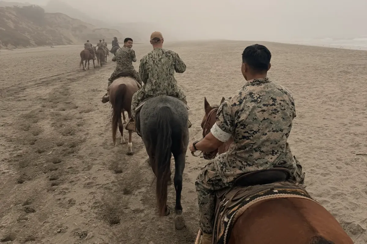 Riders in Navy and Marine Corps uniforms ride in a line along a foggy beach below sandy bluffs