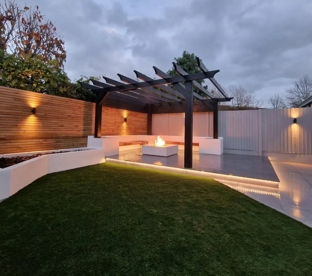 Contemporary backyard with artificial turf, custom built-in seating, wood privacy wall, and black pergola structure illuminated by soft outdoor lighting – designed by Timeless Design and Build in San Diego.
