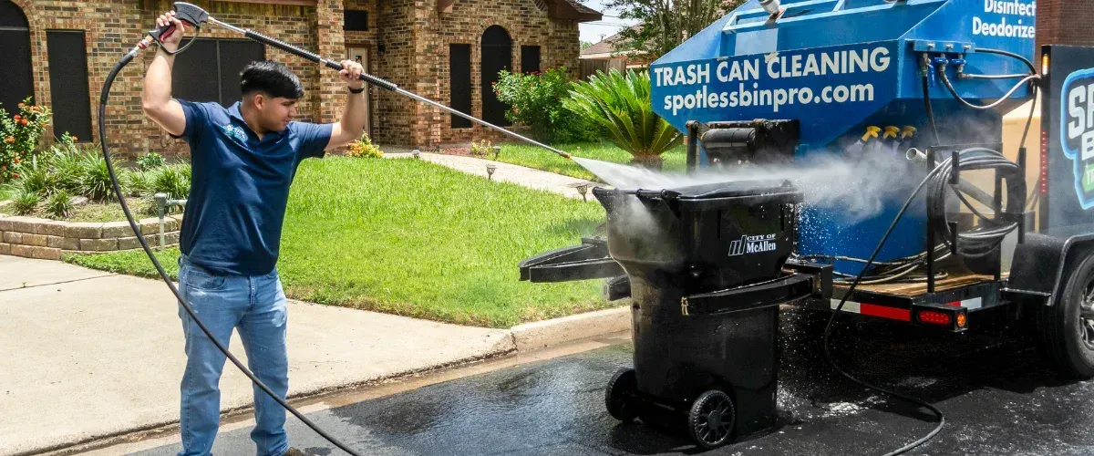 A man in a blue shirt and jeans power washes a black trash bin with a specialized cleaning machine on a sunny day.