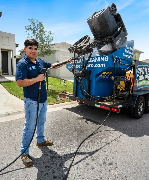A man in a navy polo and jeans power washes a black trash can on a sunny driveway.