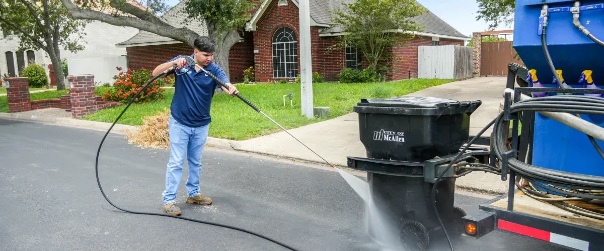 A man in blue jeans and a polo shirt power washing a black City of McAllen trash can on the street.