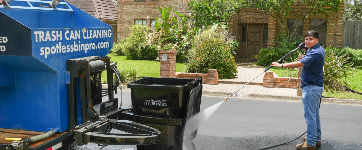 A man in a blue shirt pressure washing a black trash can on a residential street next to a blue cleaning truck.