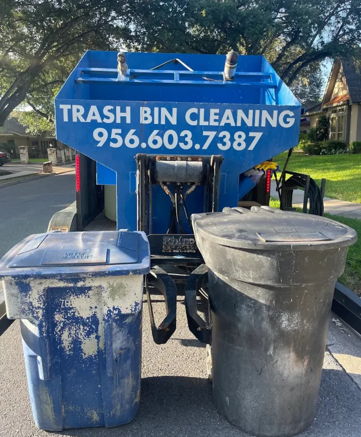 Rear view of a blue trash bin cleaning trailer with a phone number, parked with two bins on a sunny street.