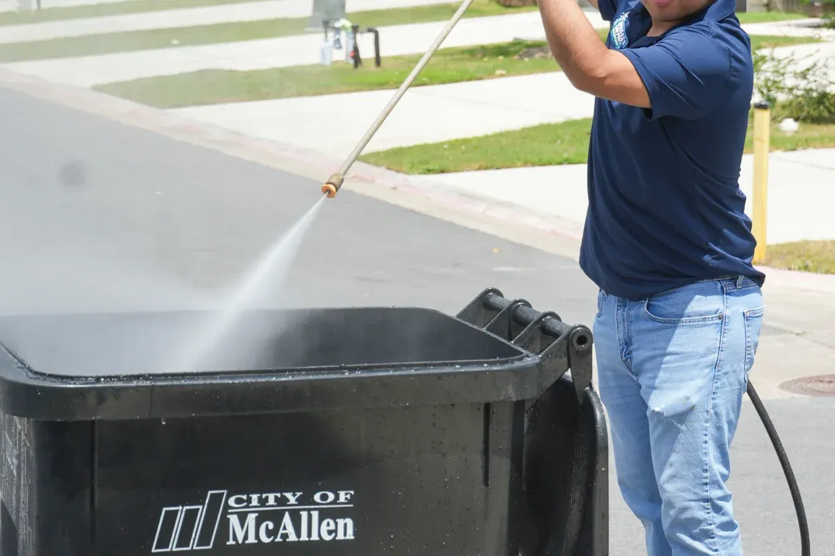 A man in a blue polo shirt and jeans pressure washes a black City of McAllen trash can on a residential street.