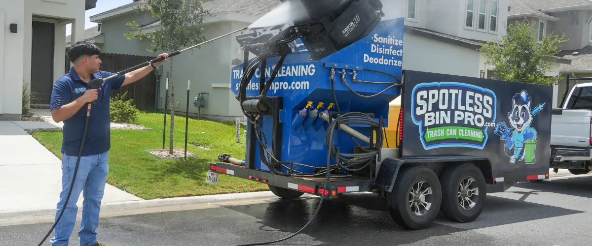 A man pressure washing a trash can lifted by a blue 'Spotless Bin Pro' trailer on a residential street.