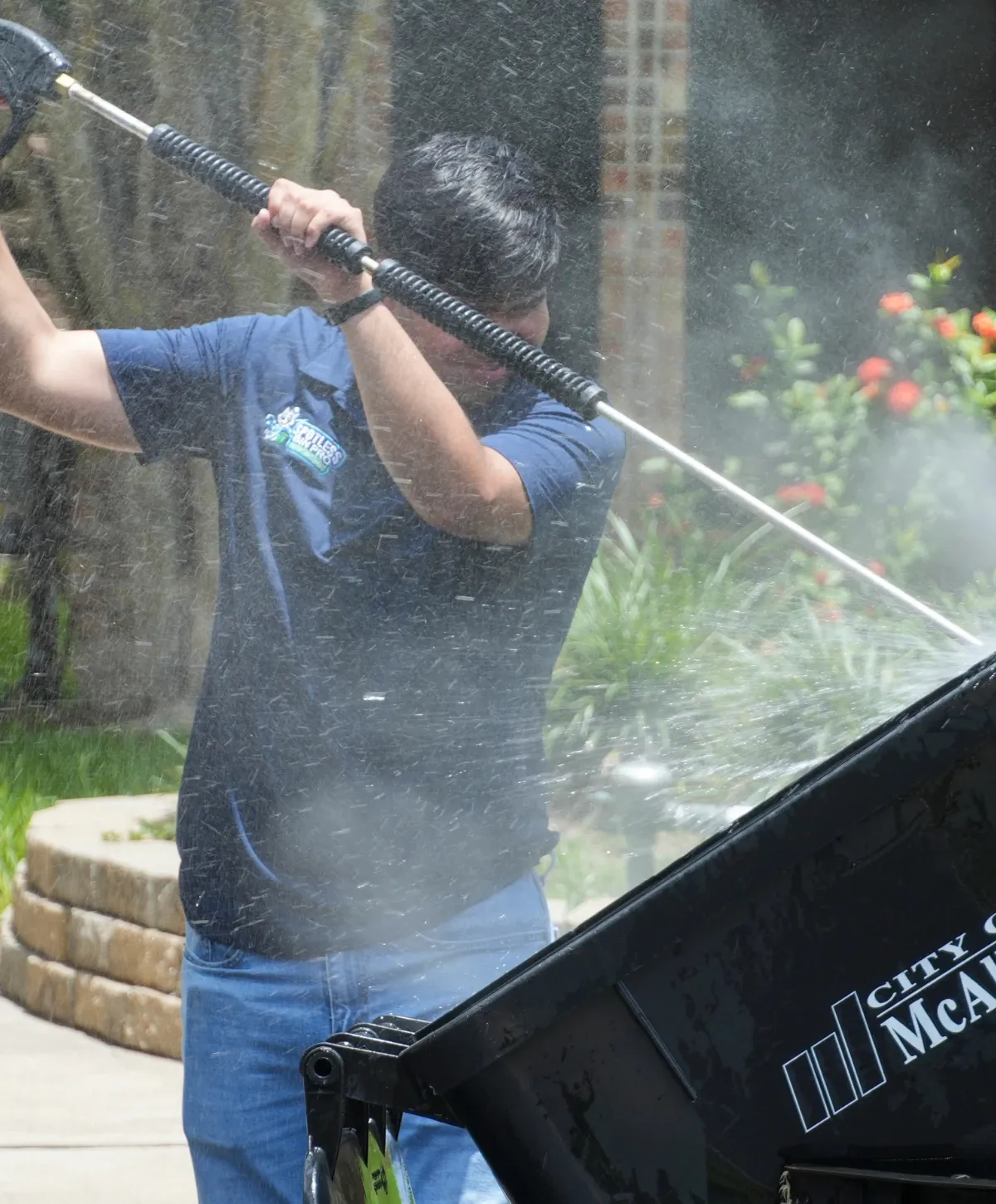 A person in a blue shirt with a logo pressure washing a black dumpster, surrounded by water spray in sunlight.