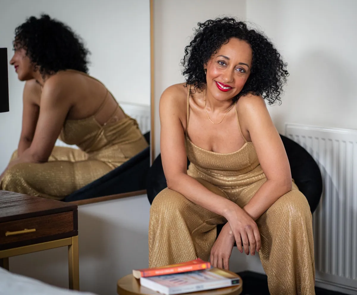 Sara Noel smiling while sitting in a gold jumpsuit, Rise Up With Sara coaching brand photo