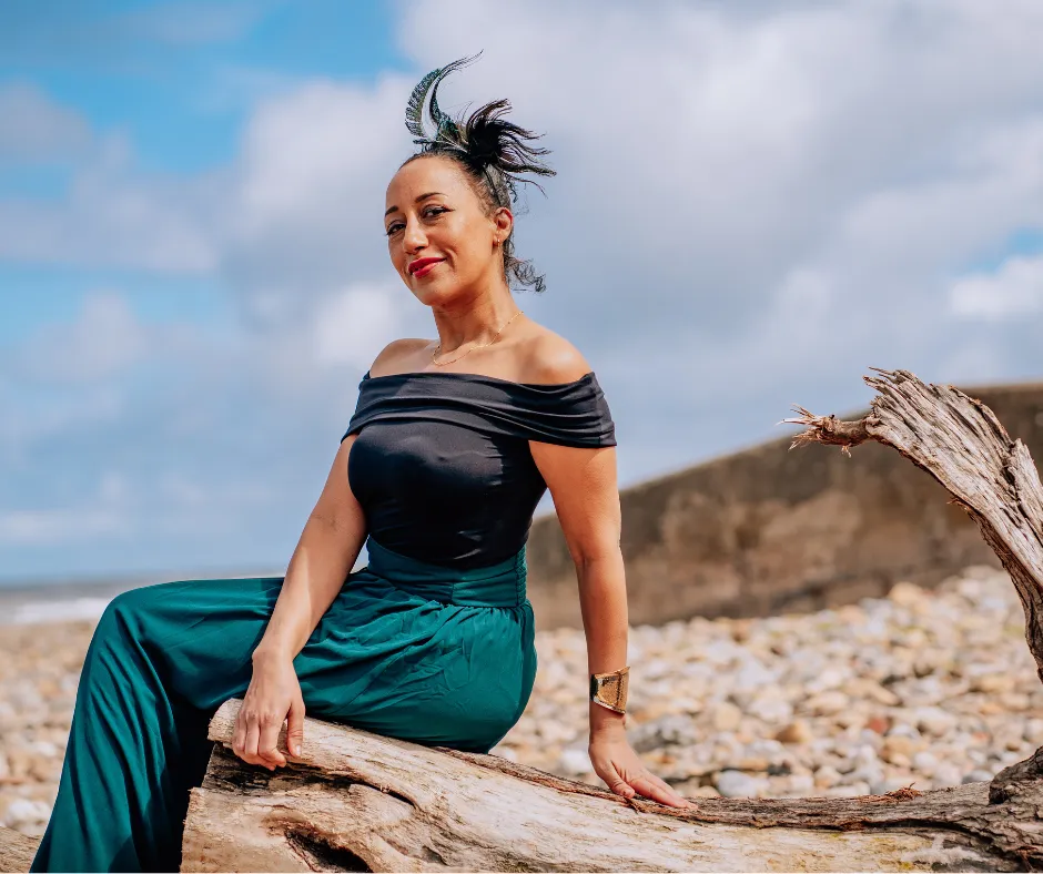 Sara Noel seated on a driftwood log by the beach in Salisbury-by-the-Sea, smiling confidently in a black off-shoulder top and teal skirt, representing balance between business success and real-life priorities.